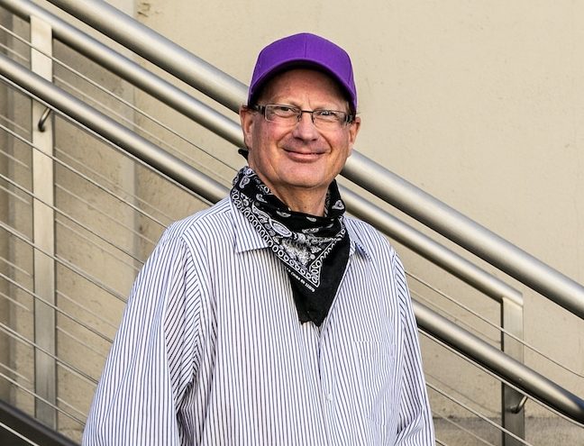 Man with purple hat and striped shirt smiling, standing against a railing