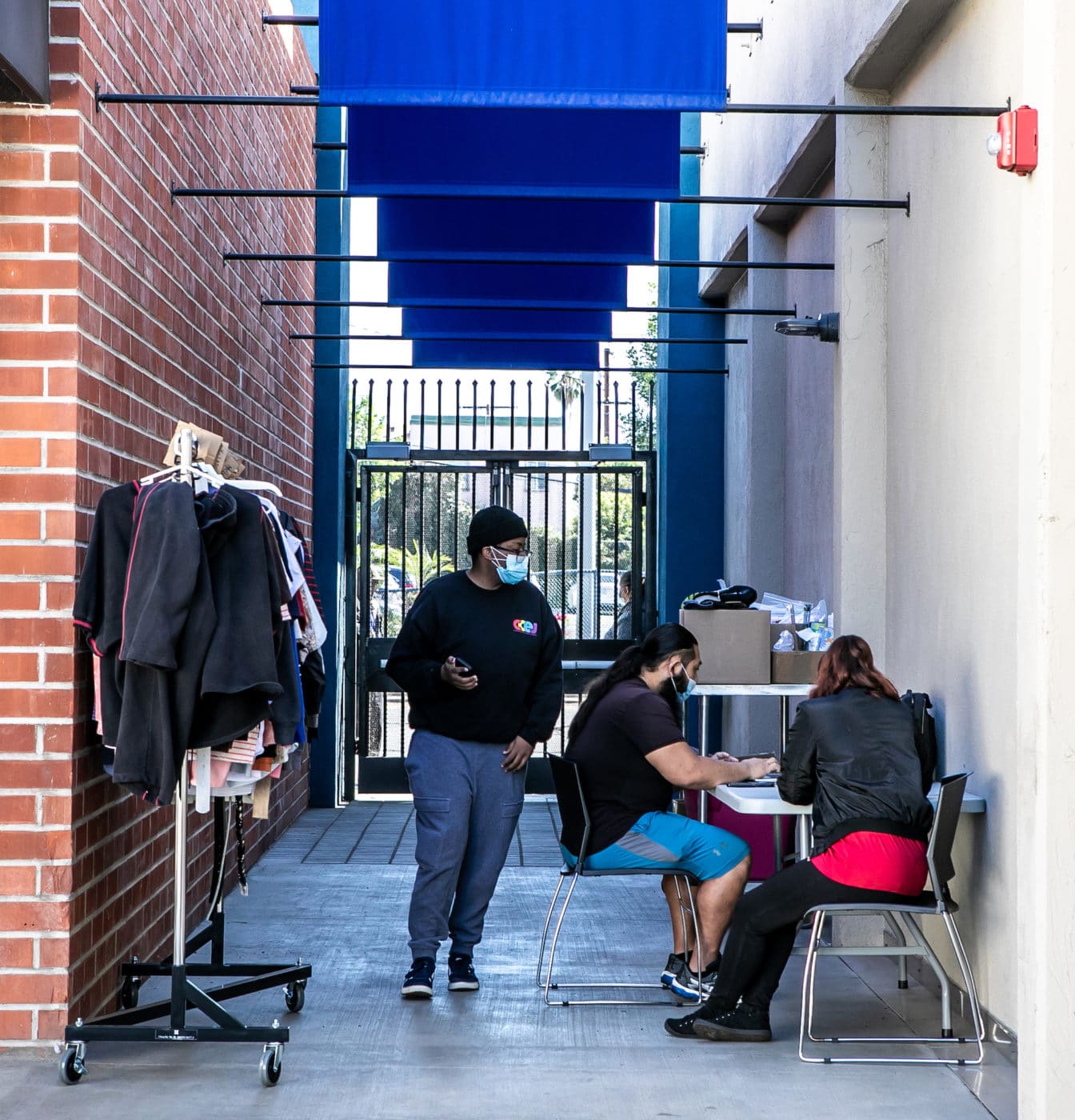 DropinOutside 2 Copy 2 seated people at a desk outside with one person standing between them and a clothing rack in an alley