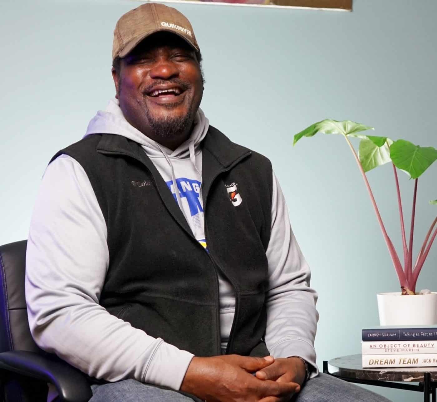 MHALA member wearing black vest and cap, sitting in front of blue wall with painting. Table with plant and books nearby.