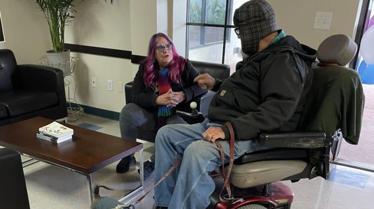 Man in wheelchair with dog talking with woman in chair. Inside, with plant and coffee table.