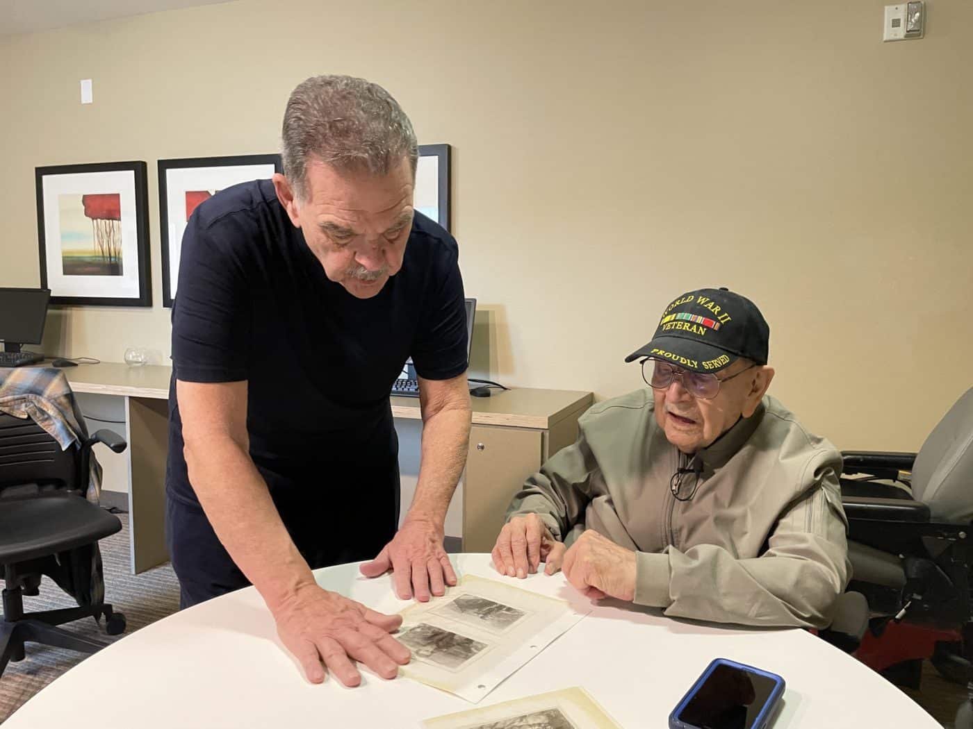 Man standing in black shirt looking at pictures with elderly man sitting at table wearing hat
