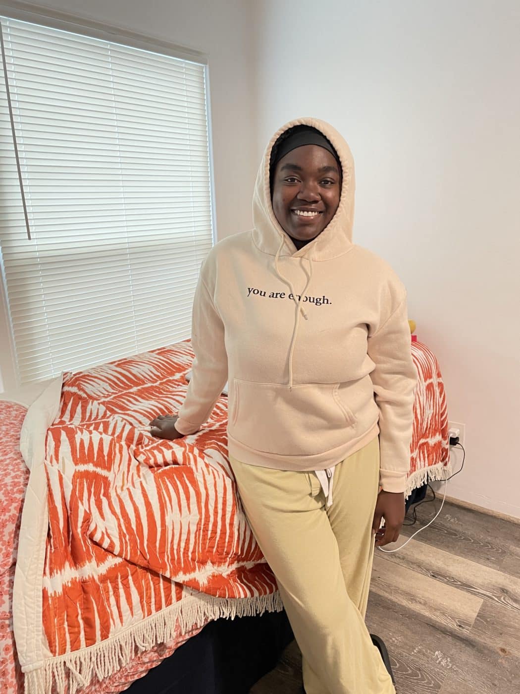 You woman in pink hoodie and sweats standing in front of bed with bright orange comforter.