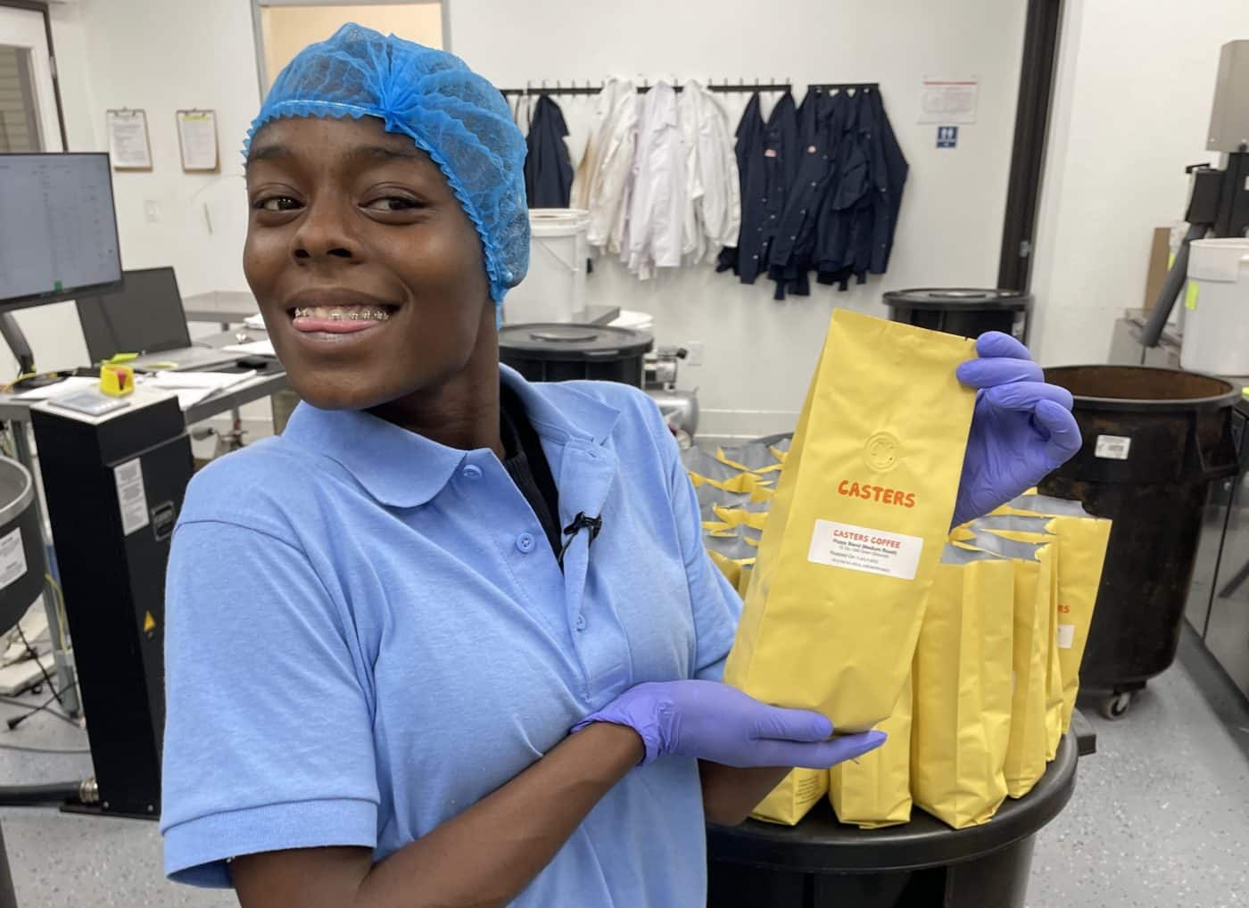 Young woman in blue hairnet and blue collared shirt holding yellow coffee bean bag in a coffee-grinding facility.