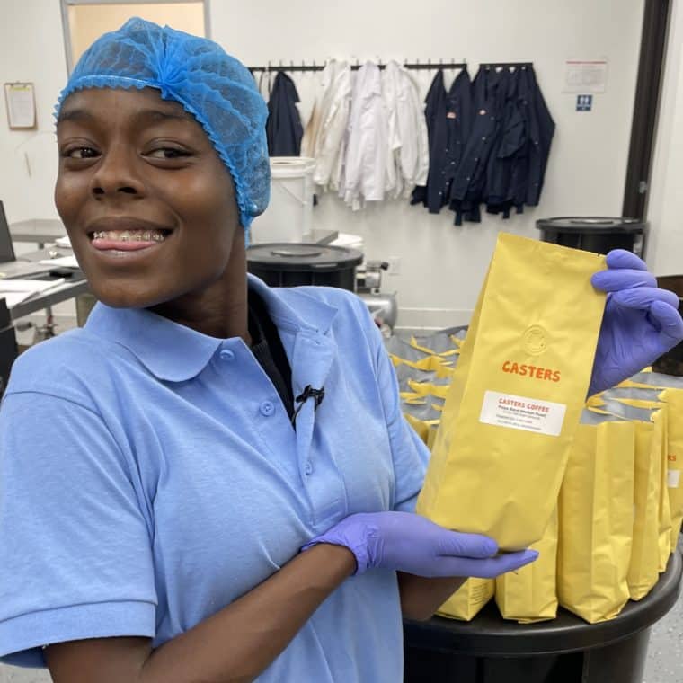 Young woman in blue hairnet and blue collared shirt holding yellow coffee bean bag in a coffee-grinding facility.