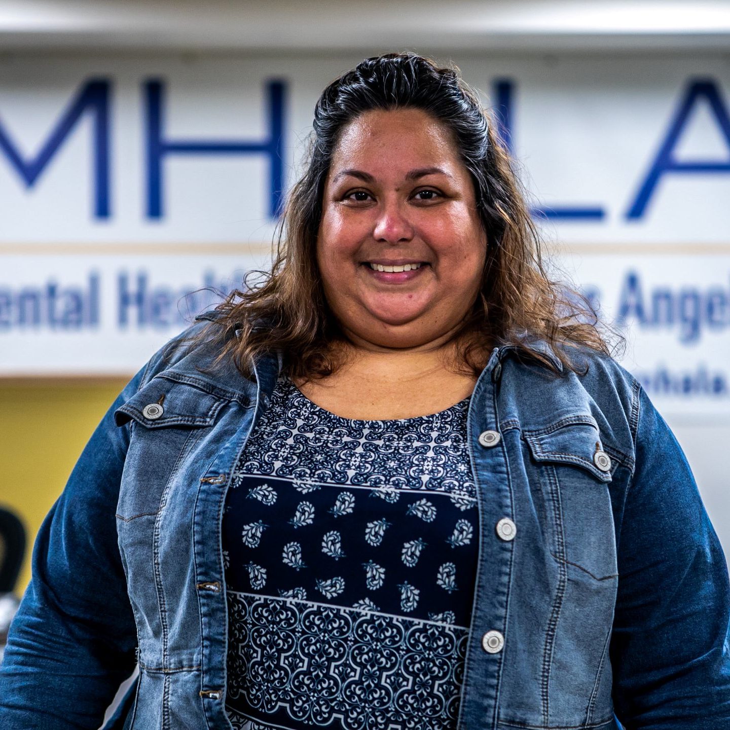 Evelyn Headshot Small MHALA member in jean jacket standing in front of MHALA banner.