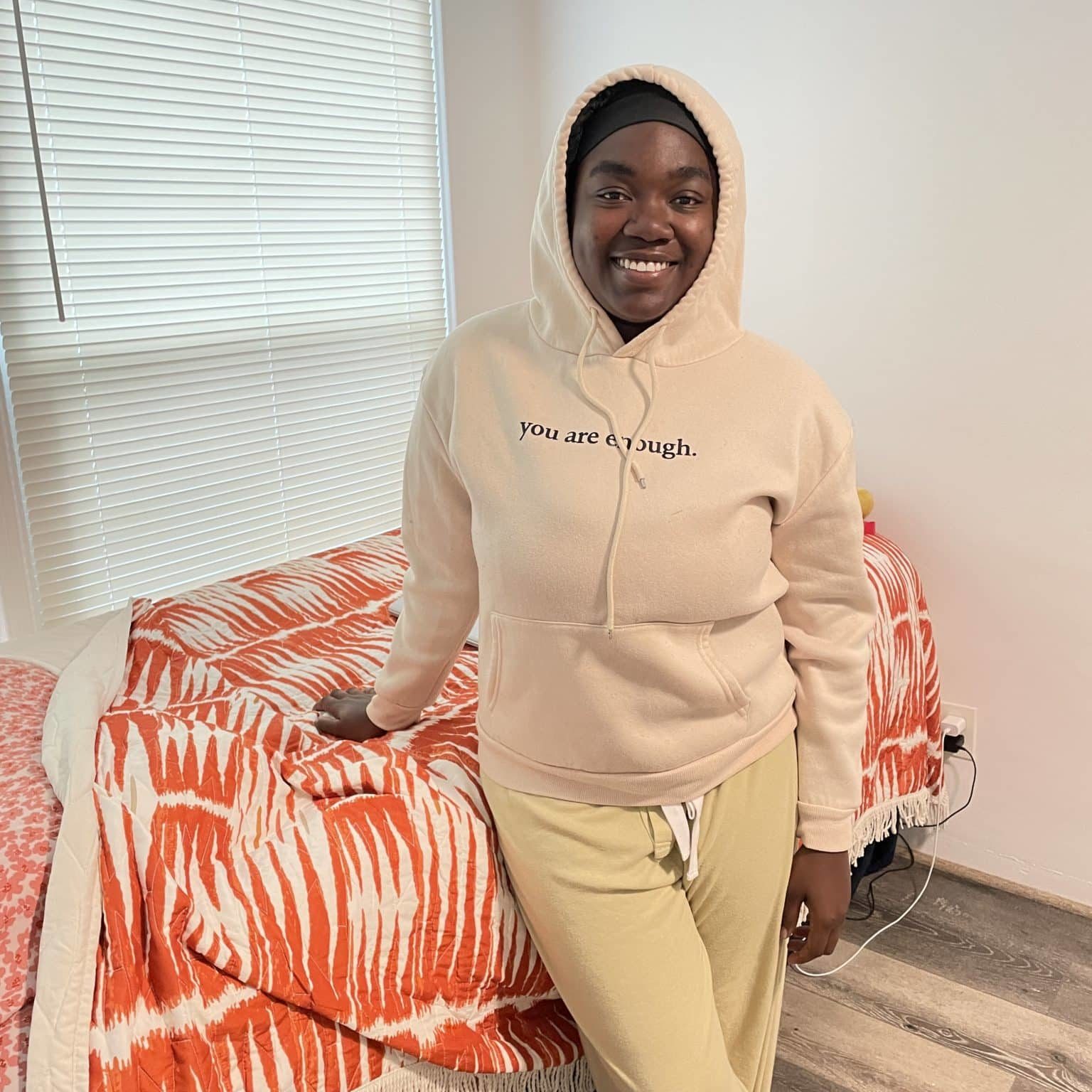 You woman in pink hoodie and sweats standing in front of bed with bright orange comforter.