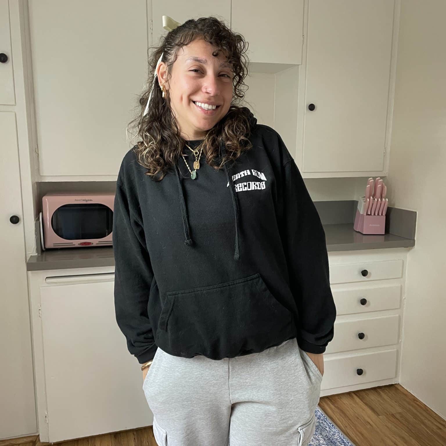 Amaya Young woman in black hoodie and gray sweatpants stands in kitchen with white cabinets and pink appliances.