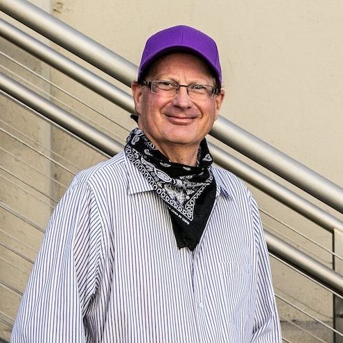 Man with purple hat and striped shirt smiling, standing against a railing