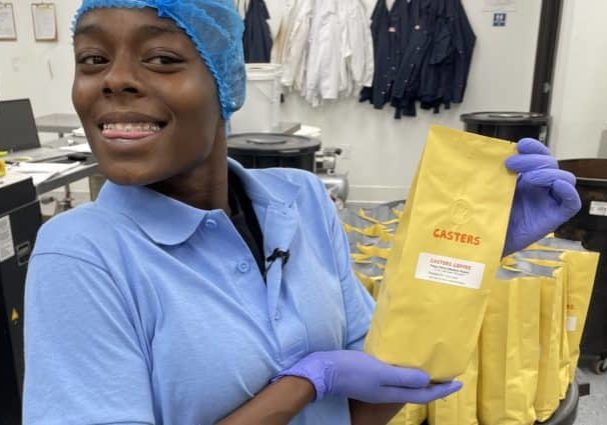 Young woman in blue hairnet and blue collared shirt holding yellow coffee bean bag in a coffee-grinding facility.