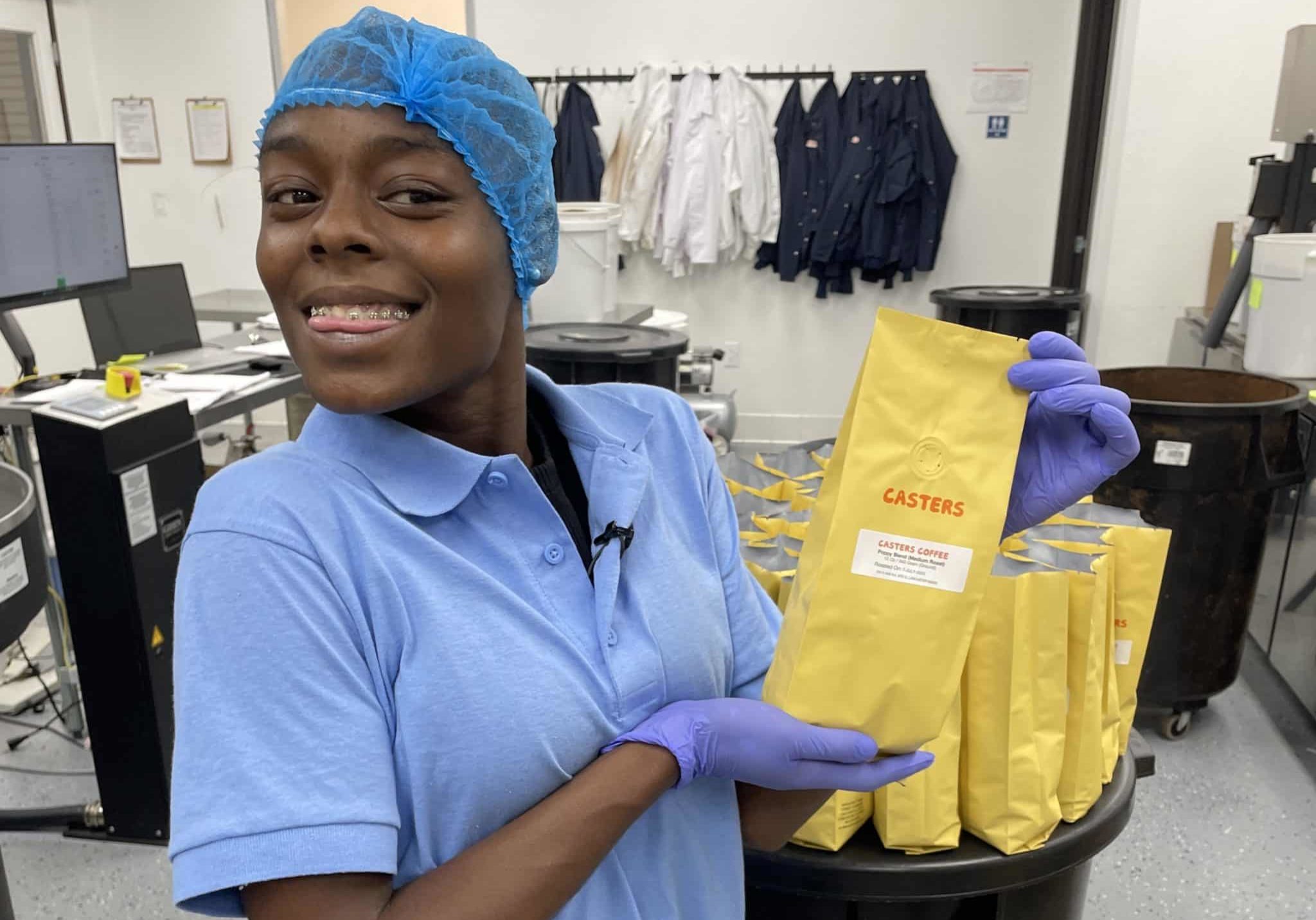 Young woman in blue hairnet and blue collared shirt holding yellow coffee bean bag in a coffee-grinding facility.